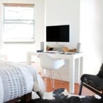 Chambre d'étudiant meublée avec lit à motifs architecturaux, bureau blanc et iMac, chaises design, tapis peau de vache, fenêtres stores bois, lumière vive.