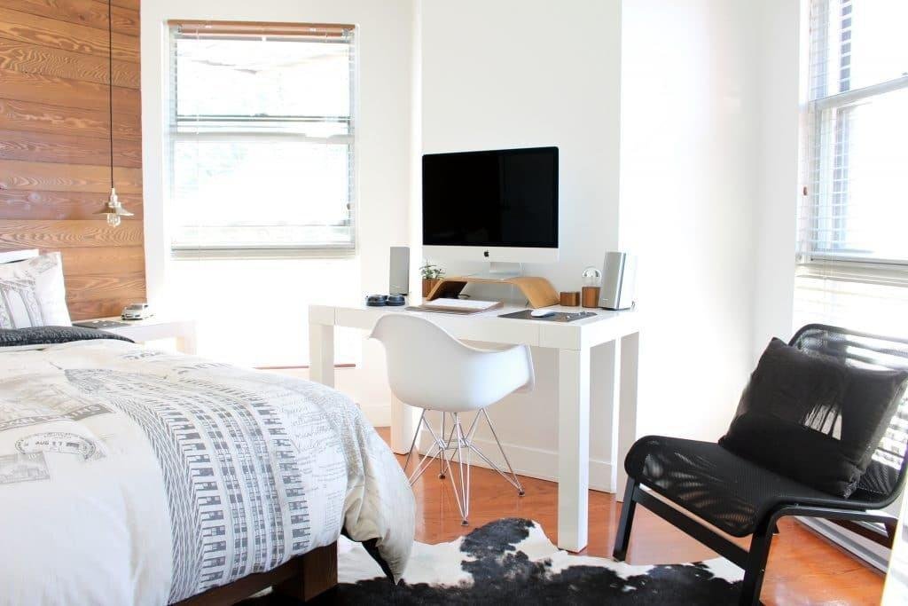 Chambre d'étudiant meublée avec lit à motifs architecturaux, bureau blanc et iMac, chaises design, tapis peau de vache, fenêtres stores bois, lumière vive.